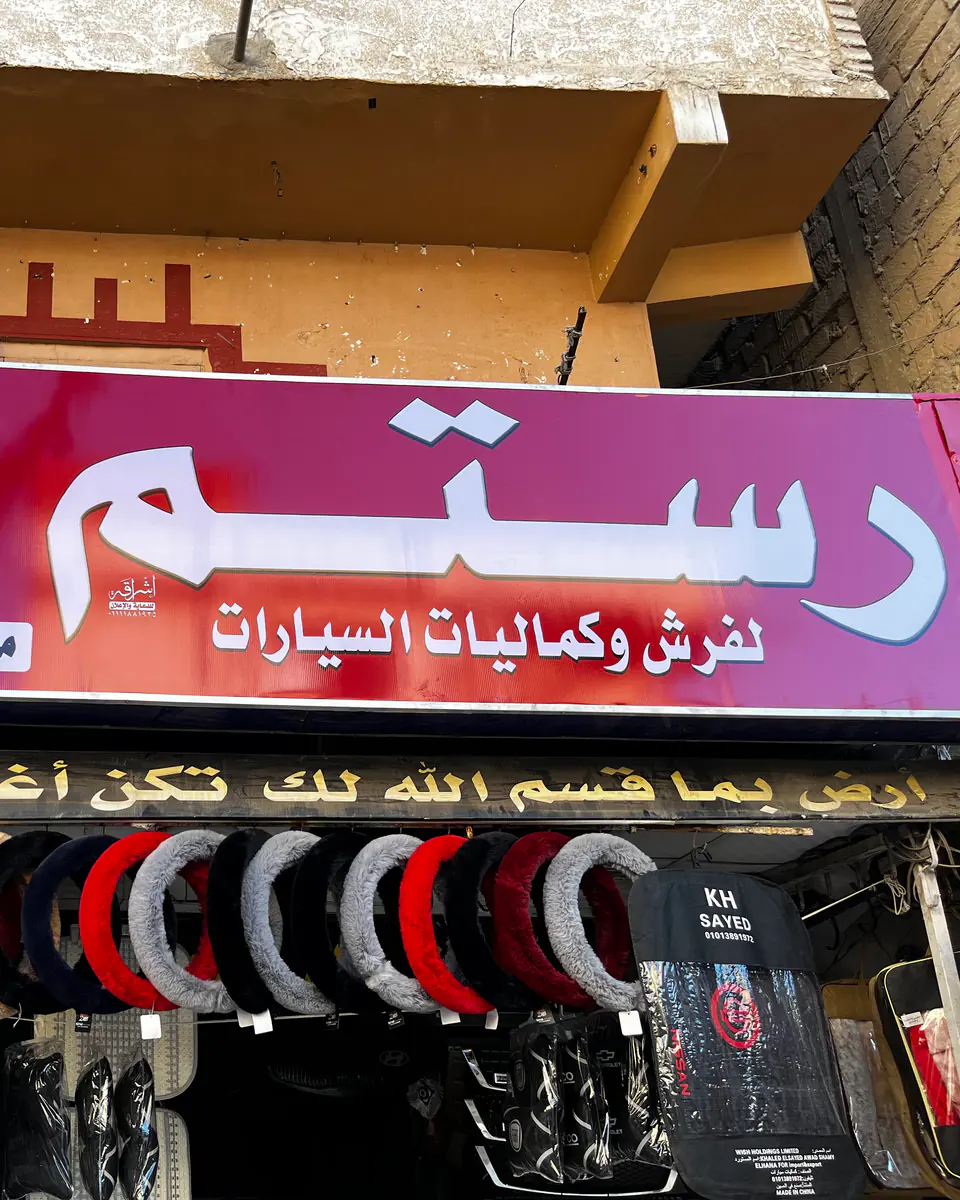 واجهة محل رستم: لافتة «رستم لفرش وكماليات السيارات» فوق أغطية دركسيون معلّقة