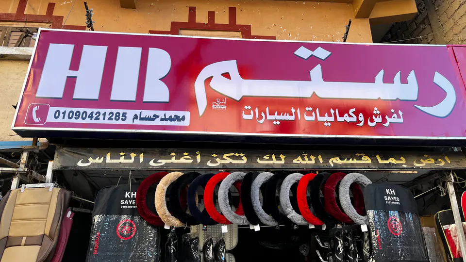 واجهة محل رستم: لافتة «رستم لفرش وكماليات السيارات» فوق أغطية دركسيون معلّقة